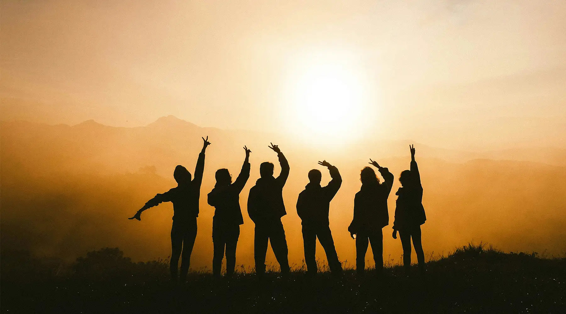 Silhouettes of a joyful group of friends with raised arms celebrating at sunset, set against a hazy golden backdrop with distant mountains, capturing the essence of freedom and adventure.