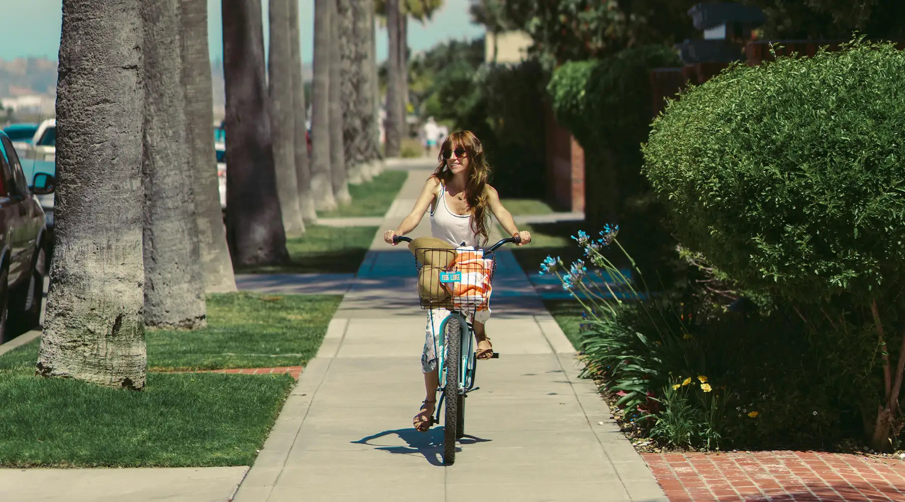 A woman rides a bicycle along a palm tree-lined sidewalk on a sunny day, smiling and wearing sunglasses. The scene is bright and cheerful with lush greenery and tall palm trees.