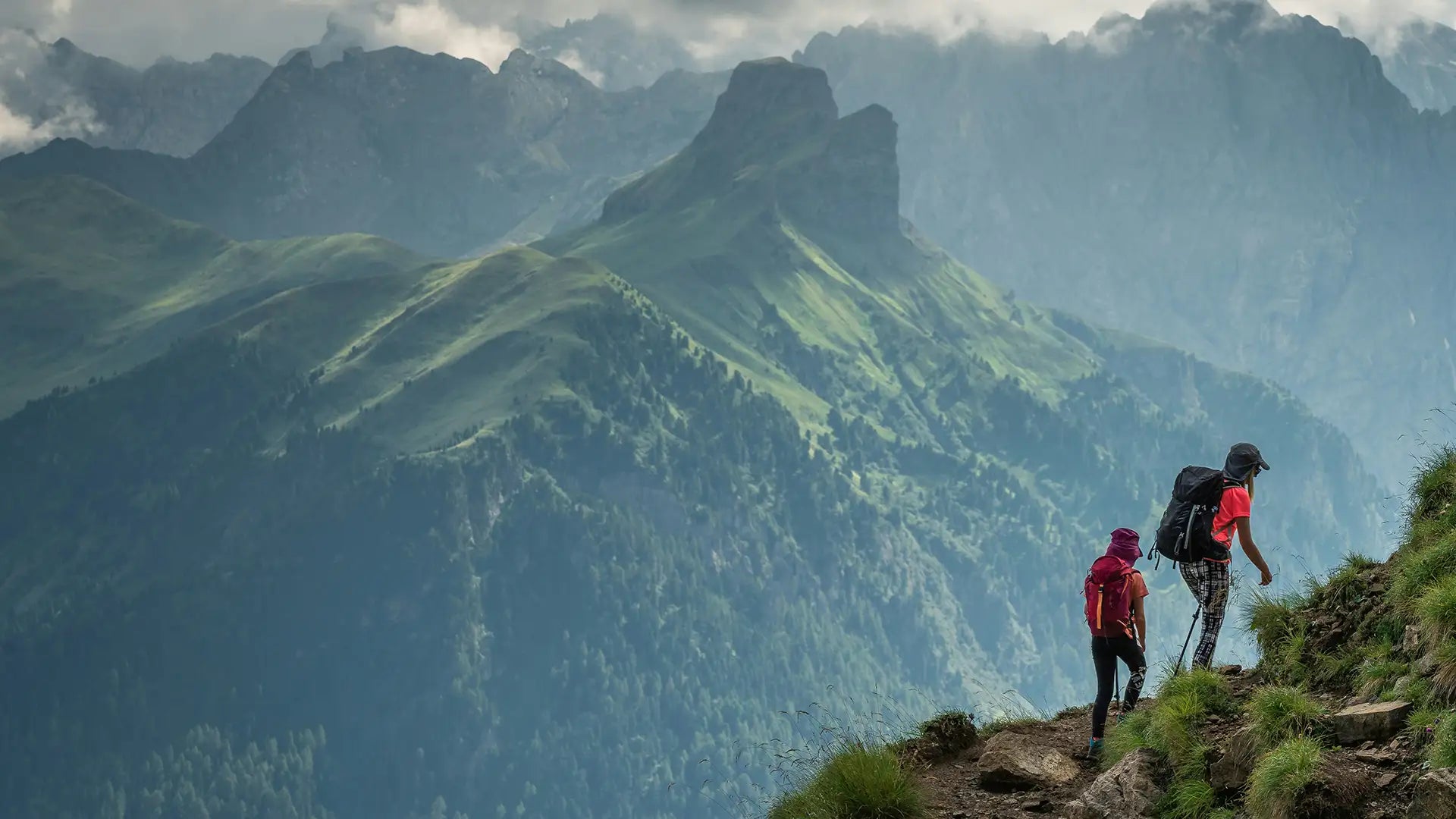 Two hikers with backpacks ascend a narrow mountain trail, surrounded by lush green slopes and dramatic cloud-covered peaks, showcasing the grandeur of alpine adventures.