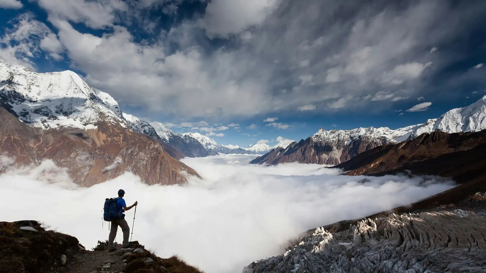 Hiker standing on a mountain trail above a vast sea of clouds, with snow-capped peaks in the distance under a dramatic sky. This stunning scene showcases the grandeur of high-altitude trekking, emphasizing the isolation and beauty accessible only to those who venture into the wilderness. The image captures the expansive views and the adventurous spirit of mountain hiking.