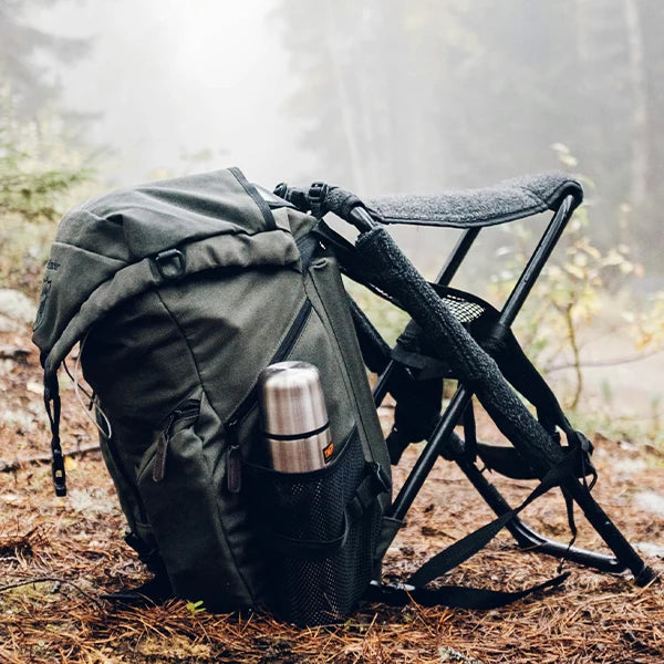 An army green hiking backpack with a built-in foldable chair, equipped with walking sticks and a stainless steel thermos, set in a misty forest environment.