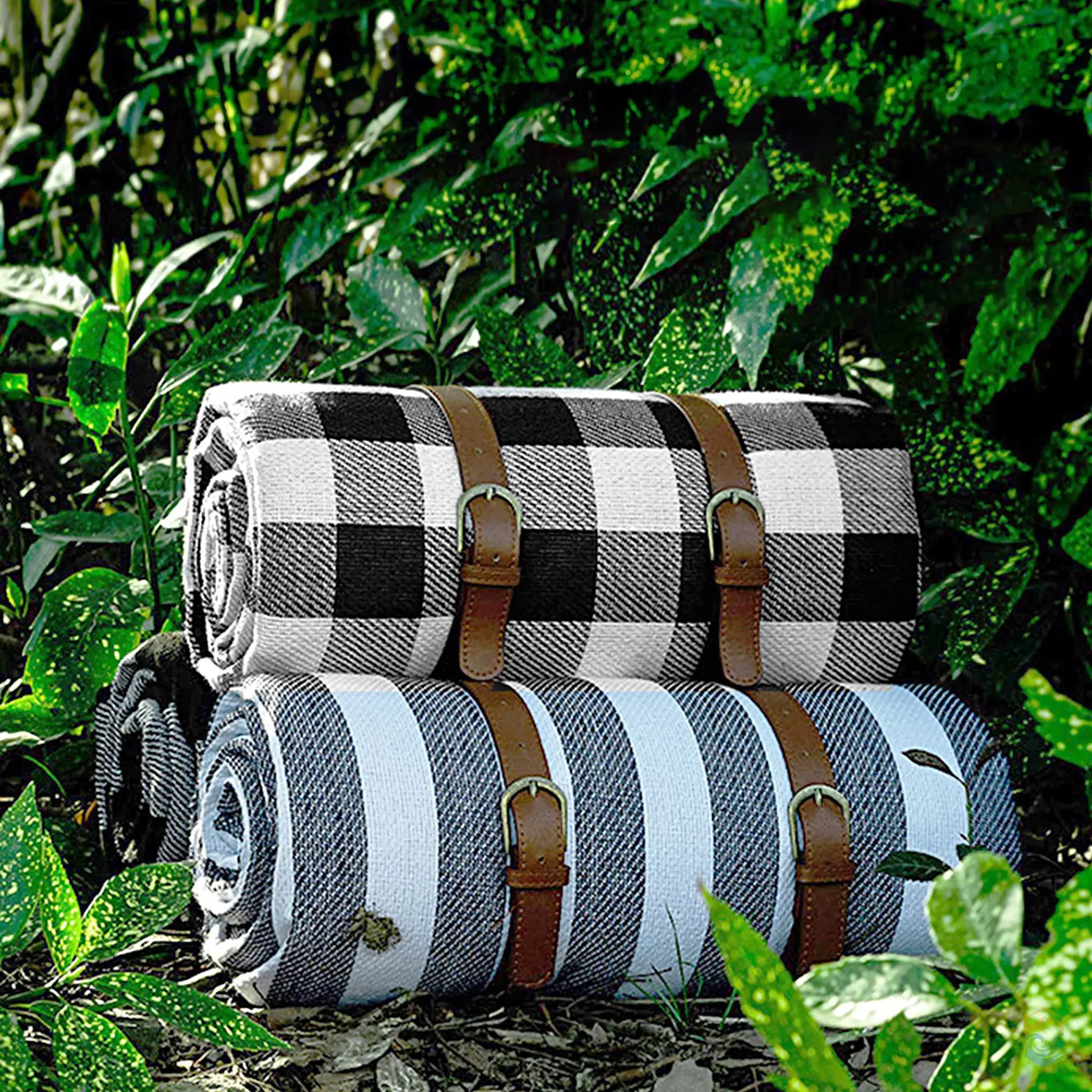 Three rolled-up checkered picnic blankets, one black and white and the other blue and white, with brown leather straps, displayed outdoors in a lush green setting.