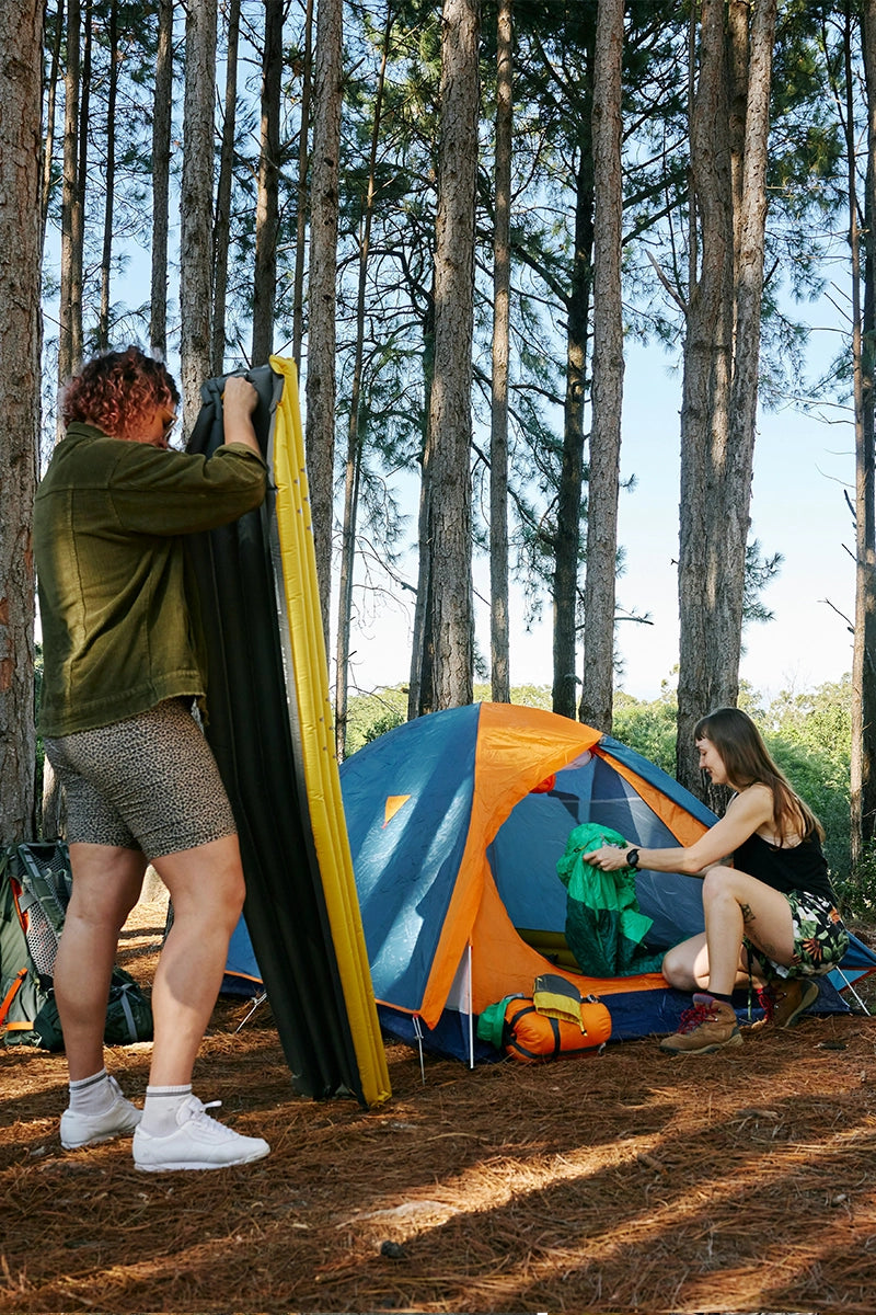 Two campers setting up a tent in a pine forest, preparing for a camping adventure amidst tall trees and serene surroundings.