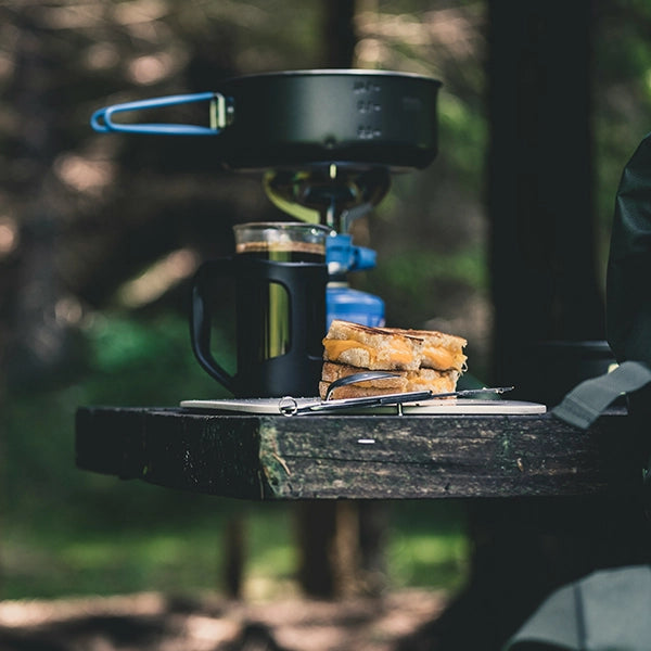 Camping setup on a wooden table featuring a sandwich, a black mug, and a portable stove with a pot in an outdoor forest setting.