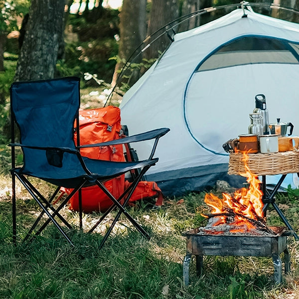 A camping scene featuring a foldout chair, a white tent, and an open fire with a cooking setup, located in a forested area.