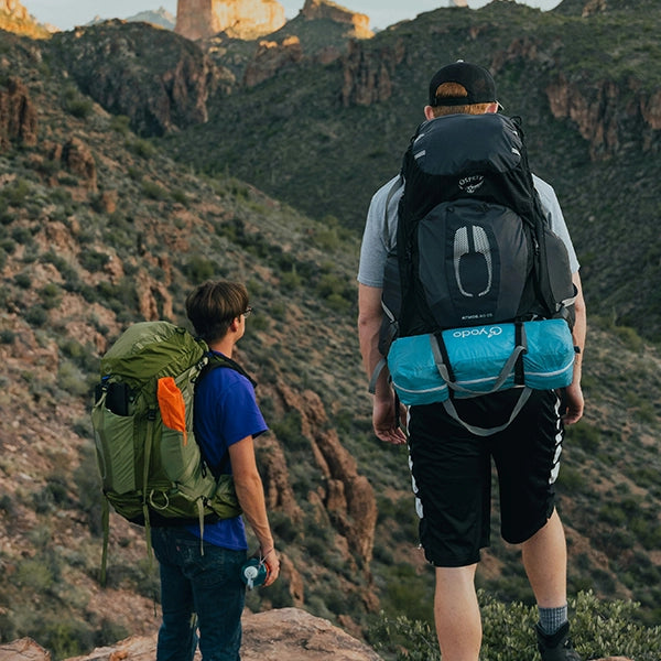 Two hikers with large camping backpacks standing on a rocky outcrop overlooking a mountainous landscape during a hiking trip.
