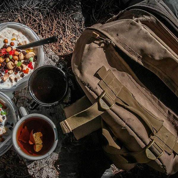 Overhead view of a camping setup with a brown canvas backpack, two cups of tea, and bowls of cereal with dried fruit on a tree stump in an outdoor setting.