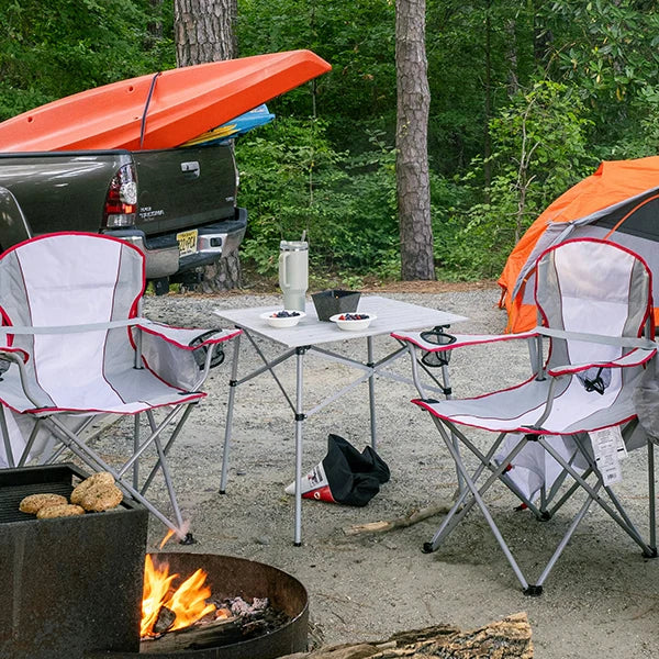 Camping setup with a table and two camping chairs, a fire pit, and a truck with kayaks in the background, situated in a forested area.