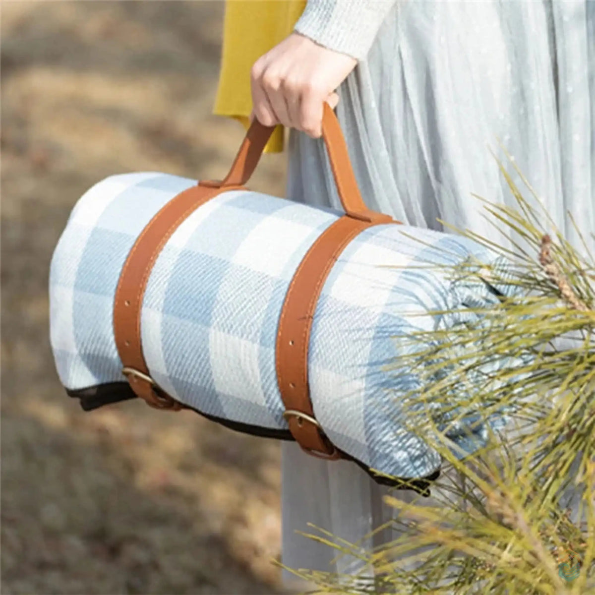 Person carrying a rolled-up blue checkered picnic blanket with brown leather straps, highlighting the blanket's portability and stylish design.