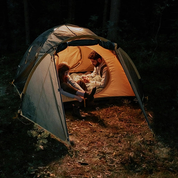 A couple sitting inside a lit camping tent at night, surrounded by a forest setting, sharing a quiet moment together.