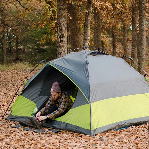 A camper tying his boots outside a half-gray, half-green camping tent, surrounded by autumn leaves and trees in a forest setting.