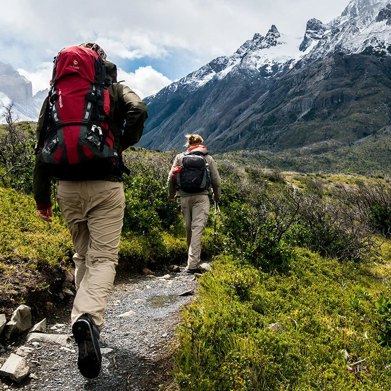 Two hikers with backpacks walking along a scenic mountain trail with snow-capped peaks in the background.
