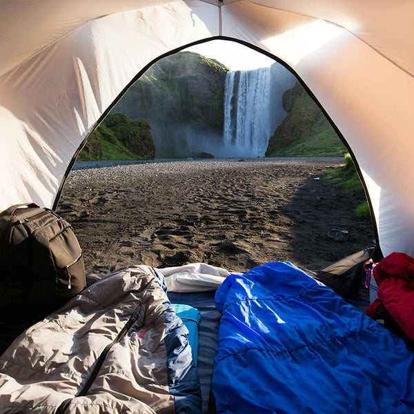 Inside view of a camping tent looking out at a stunning waterfall, with sleeping bags and camping gear arranged on the ground.