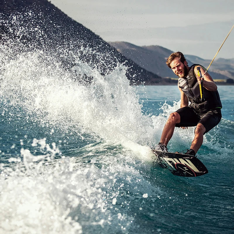 A man with a black life vest wakeboarding on a lake, creating a large splash of water, with mountains in the background and a clear sky.