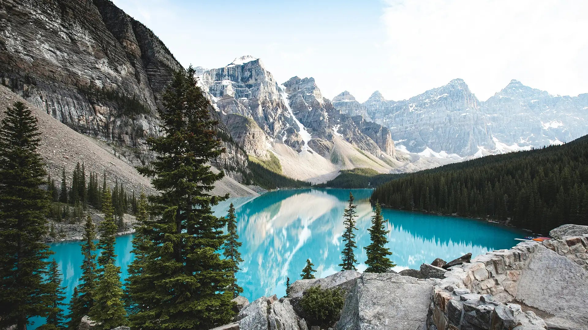 Scenic view of Moraine Lake with turquoise waters and snow-capped mountains in the background, surrounded by lush green pine trees and rocky terrain.