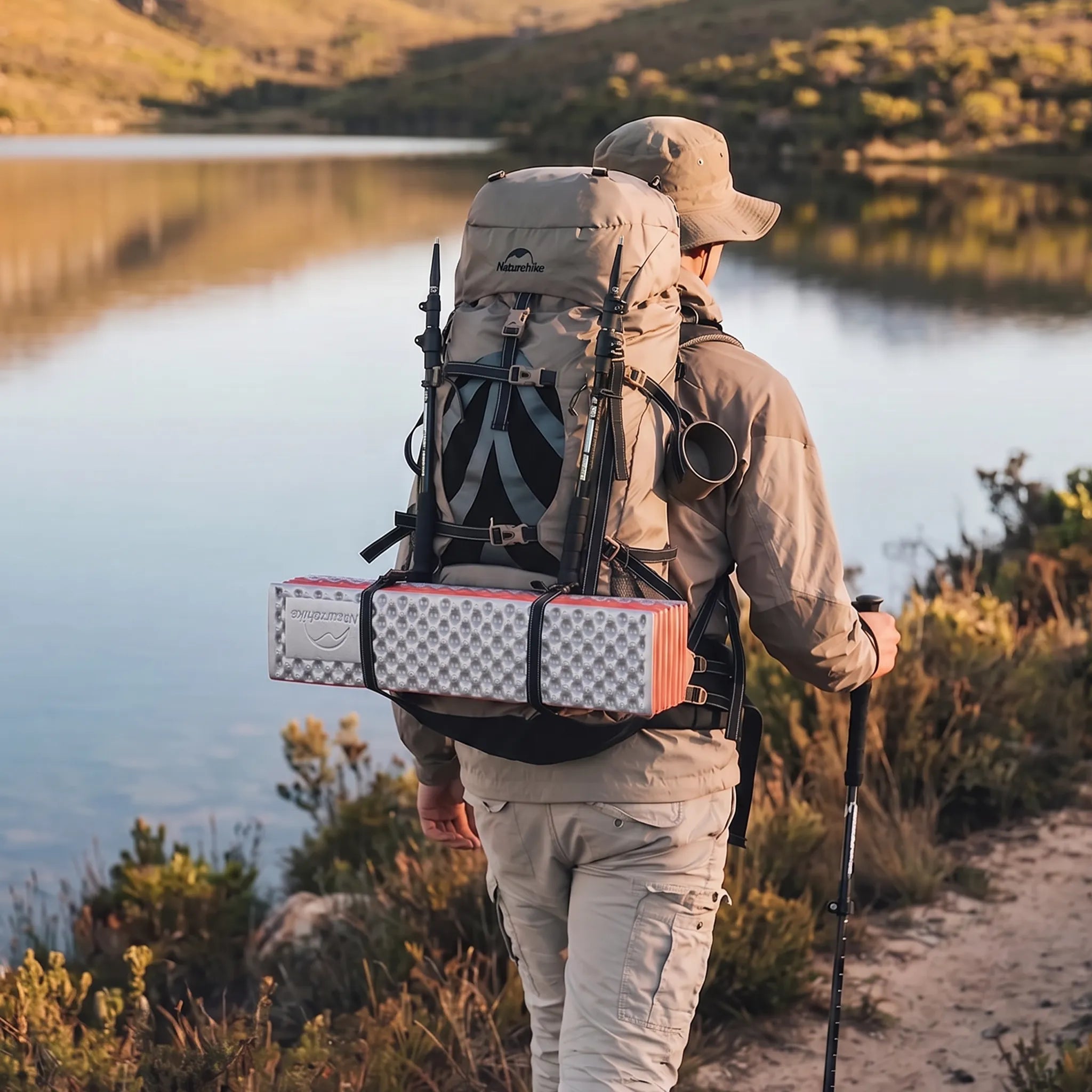 Hiker wearing the Naturehike Discovery 70L backpack while walking along a lakeside trail with trekking poles and camping gear attached.