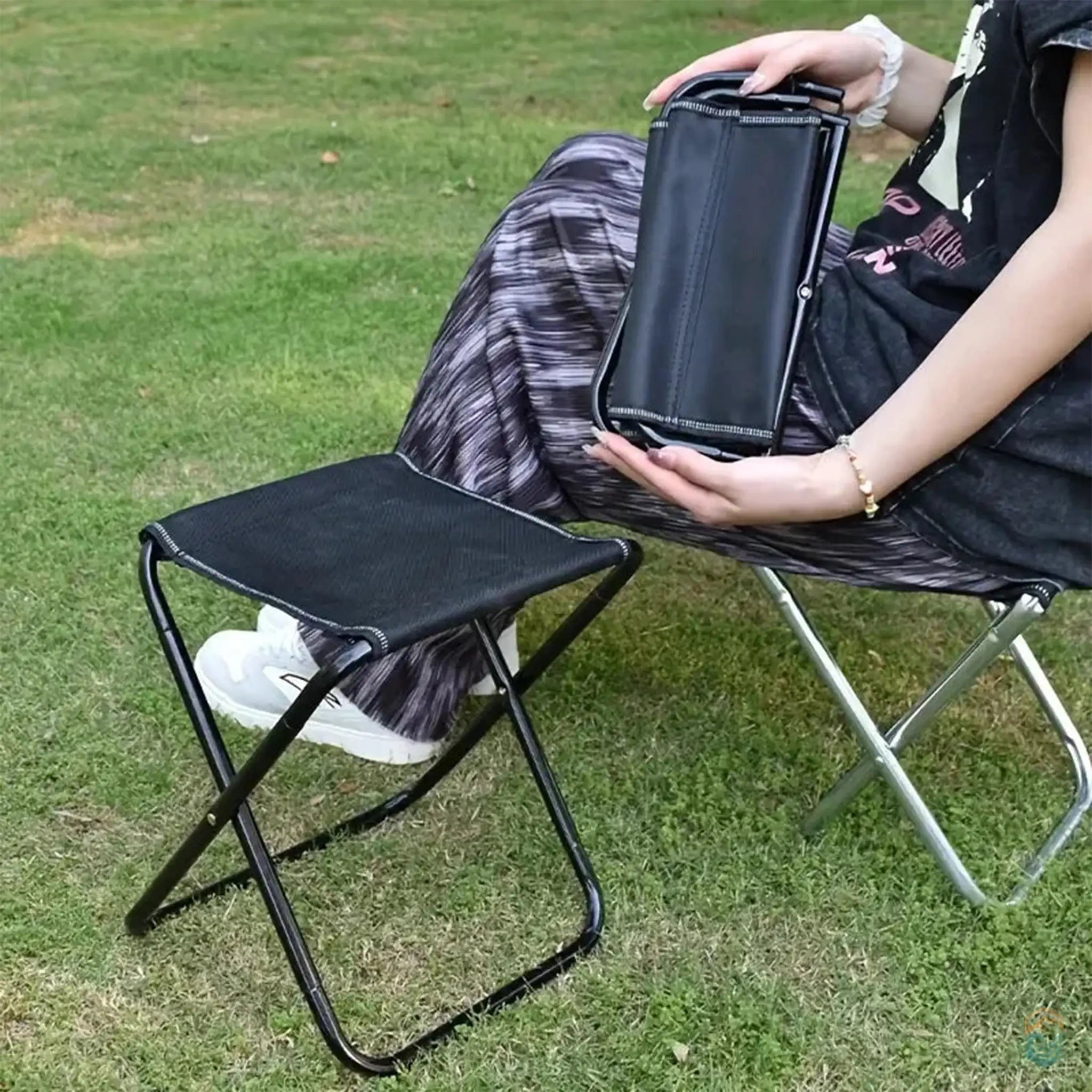 Person seated outdoors using a portable lightweight folding chair with a black seat and black legs, holding the folded version of the chair.