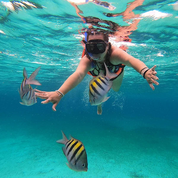 A person snorkeling underwater with colorful tropical fish, wearing a snorkel mask and life jacket in clear turquoise water.