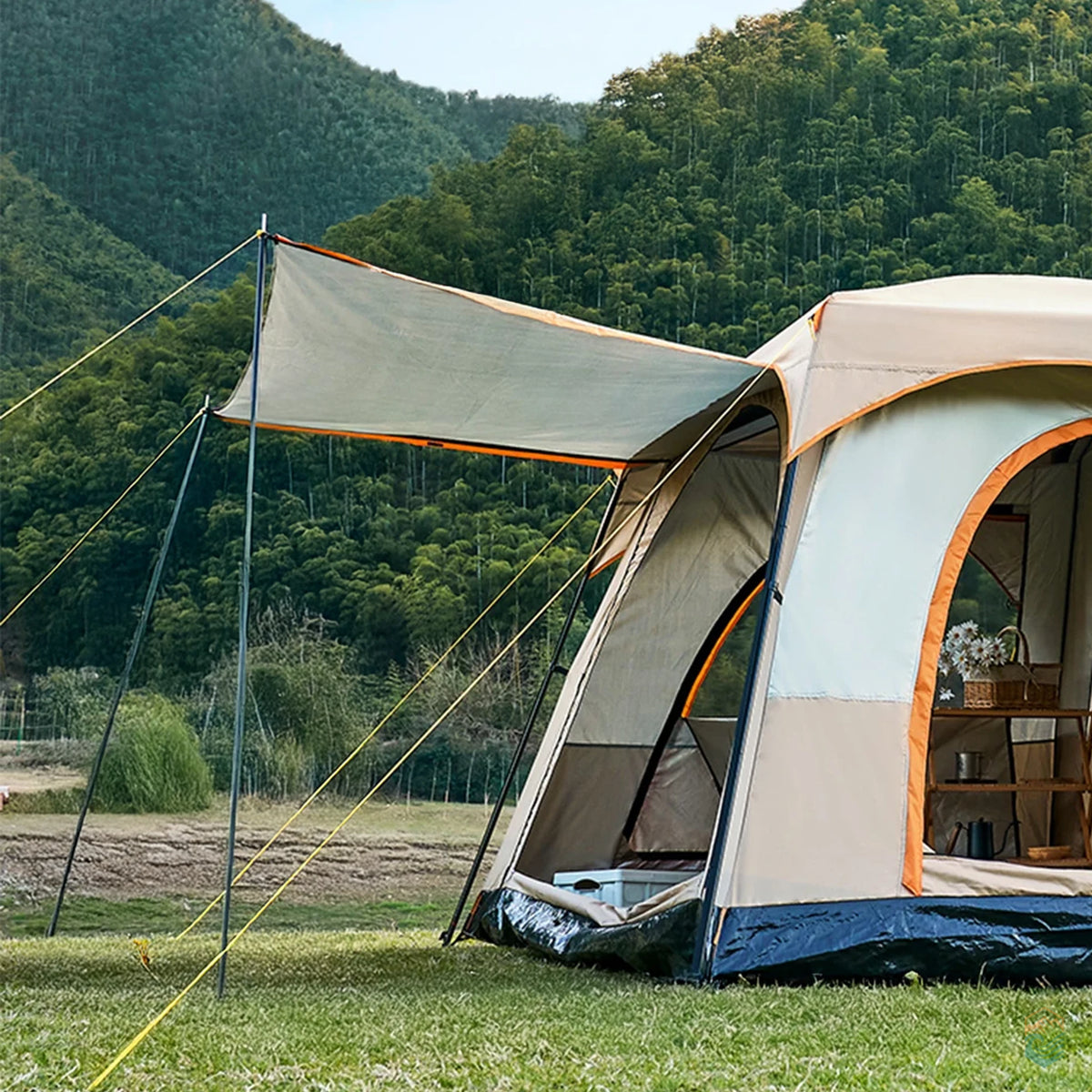 Side view of khaki Sonuto family camping tent with canopy extended, pitched on grass near a forested mountain with organized camping gear inside