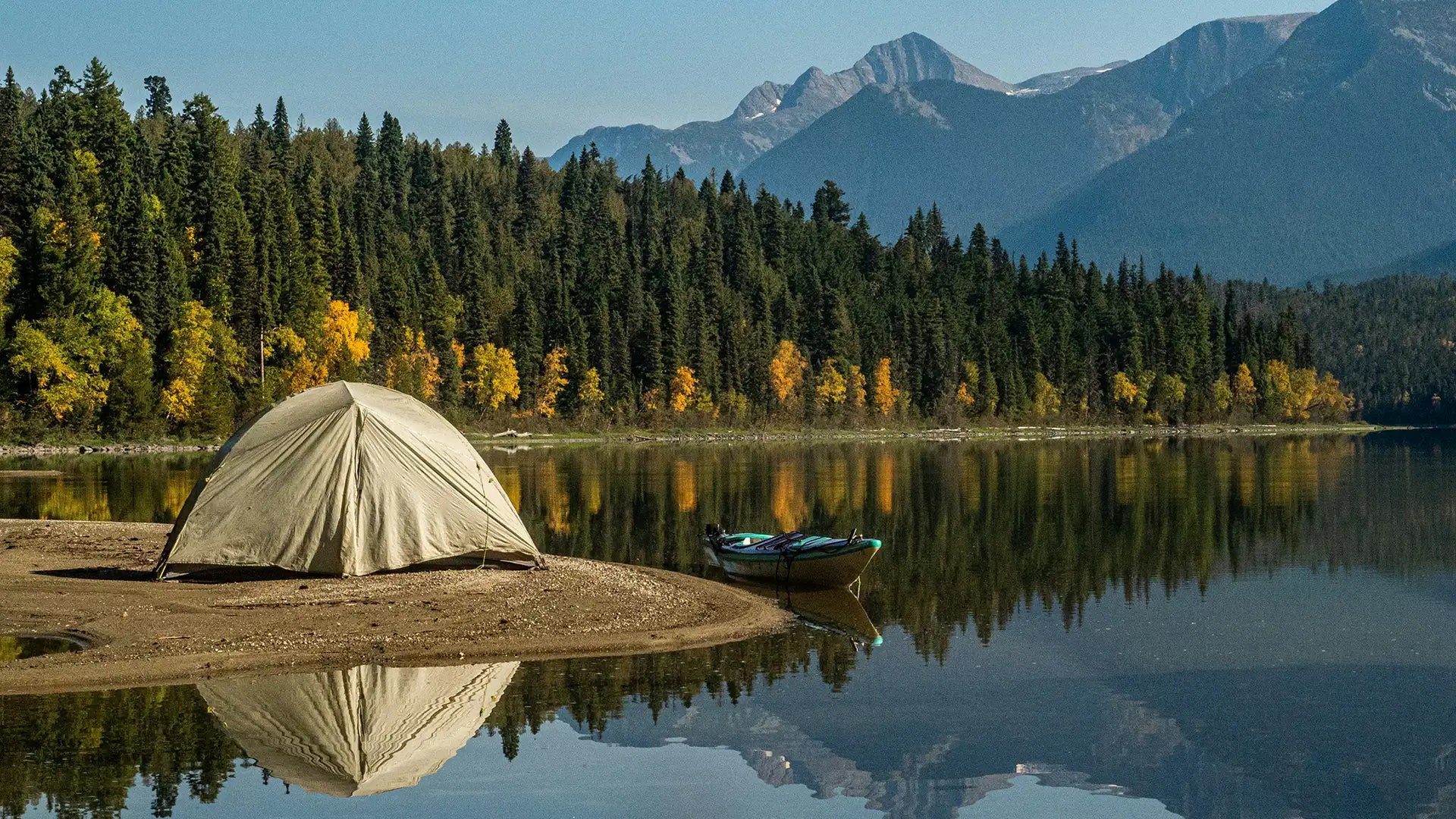 A beige tent set up on a sandy shore beside a tranquil lake, with a kayak nearby and surrounded by a dense forest of pine trees and mountains in the background. The clear water reflects the tent, trees, and mountains, creating a serene camping scene.