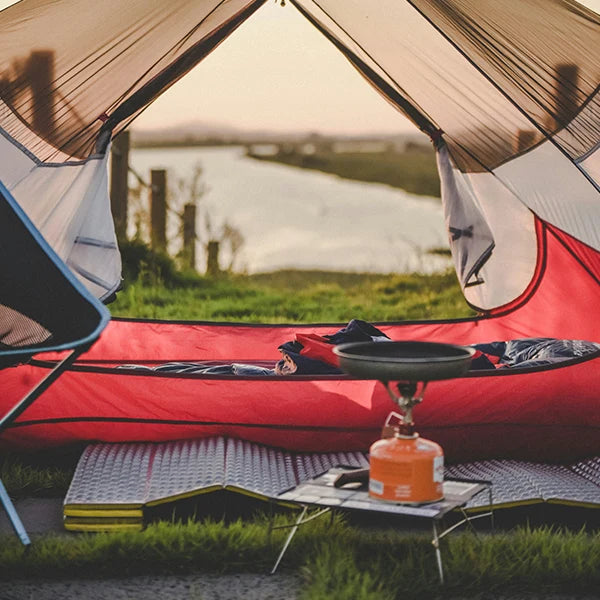View from inside a tent with camping mats, a sleeping bag, and a portable gas stove set up near a river at sunset.
