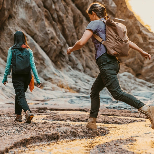 Two women hiking through a canyon, one jumping over a small stream of water, both carrying backpacks and wearing outdoor gear.