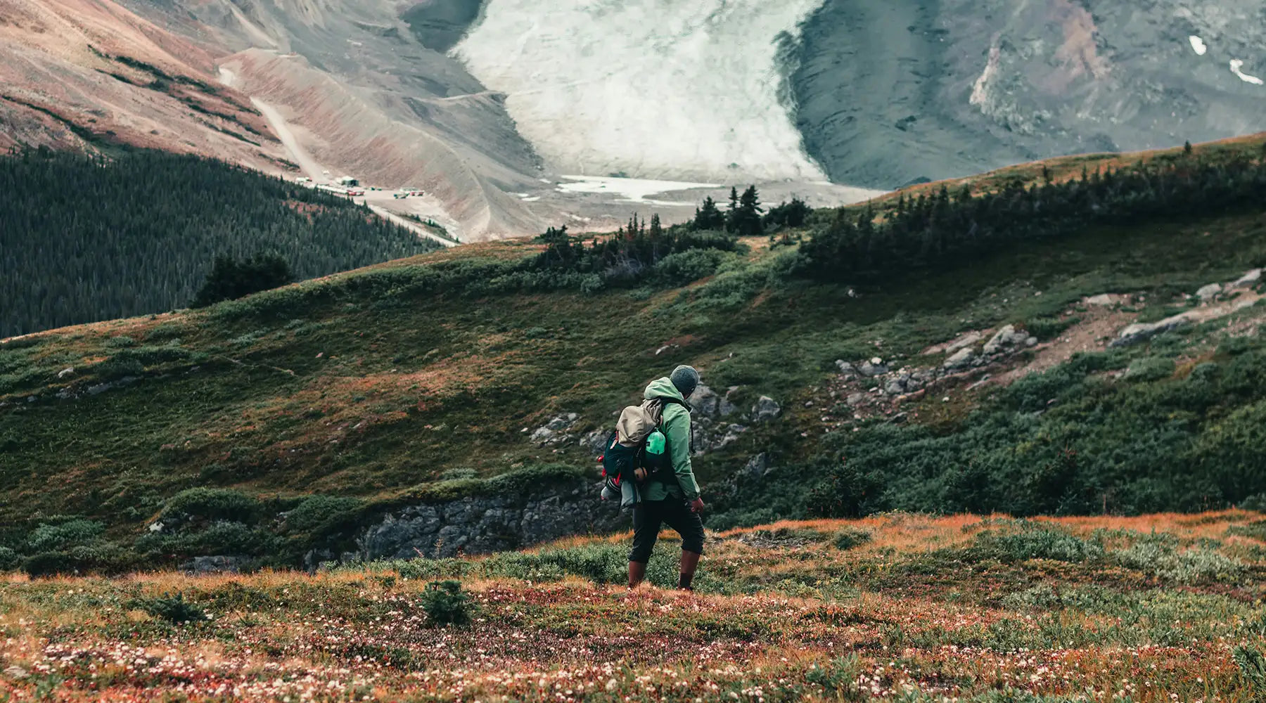 A hiker walking through a mountain valley with a glacier in the distance, surrounded by green and rocky terrain, exploring the rugged outdoor landscape.