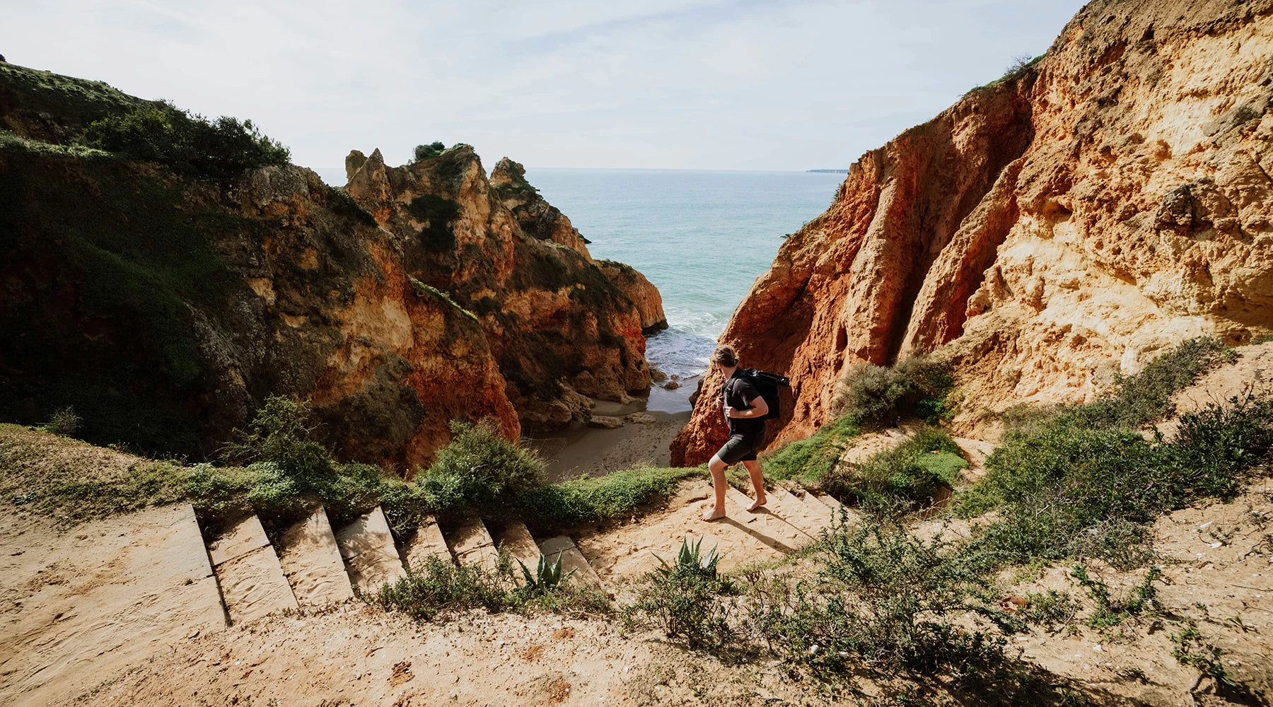 A person hiking down a staircase along a coastal cliff path with a backpack, overlooking the ocean and dramatic rock formations.