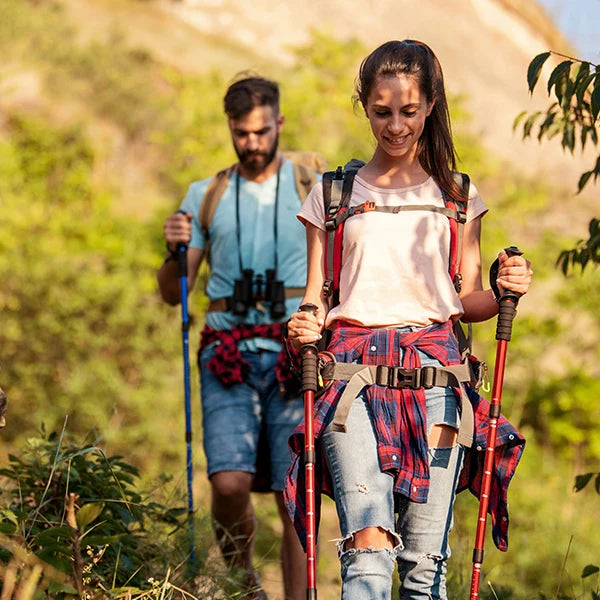 A young couple equipped with hiking gear trekking along a rugged mountain trail. The woman leads the way, smiling and enjoying the hike, while the man follows closely behind, both using trekking poles for support. Their casual yet functional attire and the lush green backdrop highlight the joy and challenge of outdoor adventures, making this image perfect for content related to hiking, nature trails, or adventure travel.