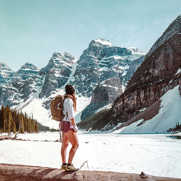 A female hiker admiring the majestic snow-covered mountain peaks from a snowy lakeside. Wearing a backpack and a cap, she gazes at the rugged landscape, capturing the essence of spring mountain adventures where snowy conditions meet clear, sunny skies. This serene moment reflects the peace and grandeur of nature, ideal for illustrating articles or promotions related to outdoor exploration and adventure tourism.