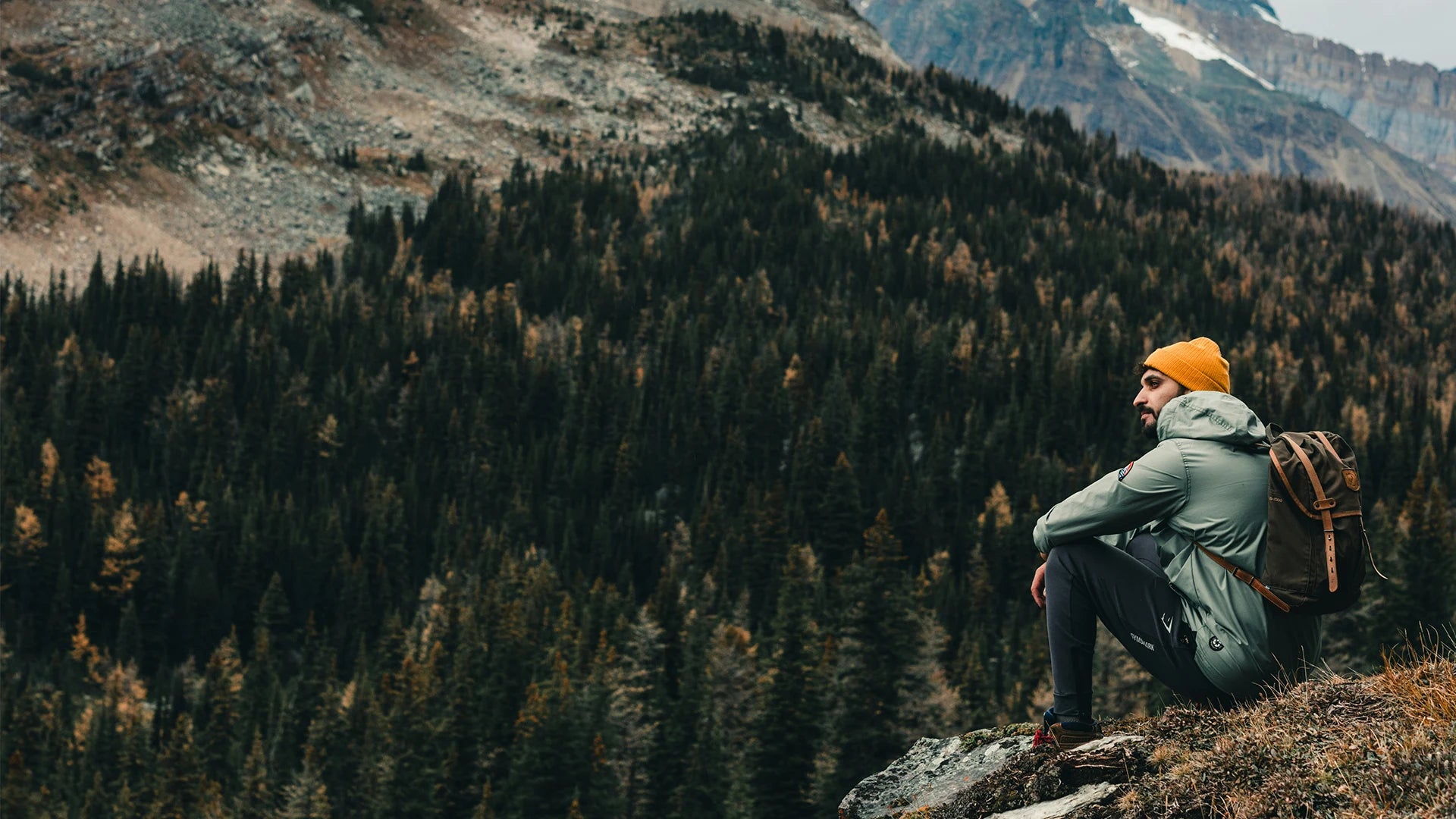 Man sitting on a rock with a camping backpack, looking out over a dense forest in the background, wearing outdoor gear and a yellow beanie.