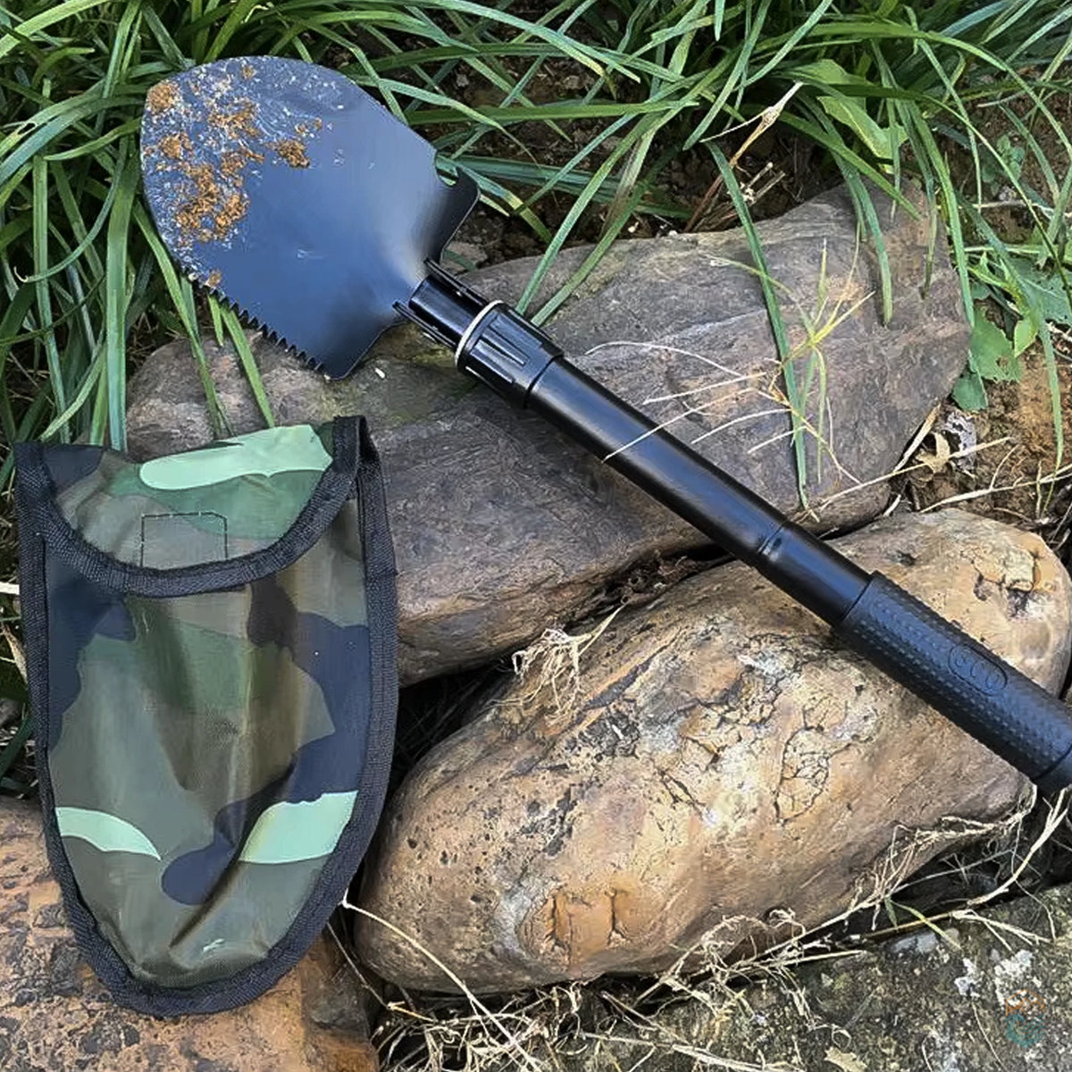 Black folding survival shovel with serrated blade and textured handle resting on rocks next to camouflage carrying pouch, shown with dirt on blade after outdoor use