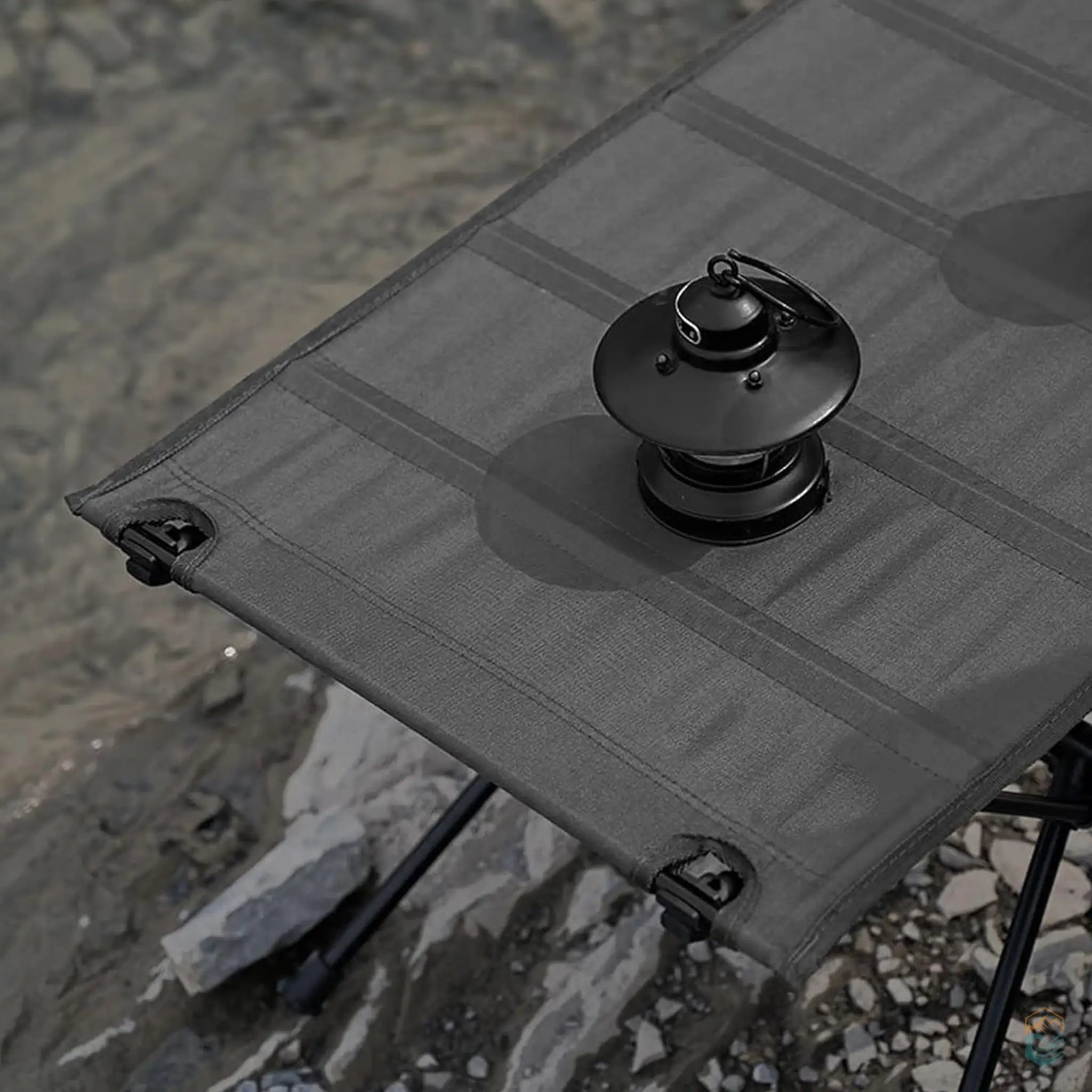 Close-up view of a black collapsible camping table with an aluminum frame, featuring an outdoor lantern on the table surface, set on rocky terrain, ideal for camping and outdoor activities.