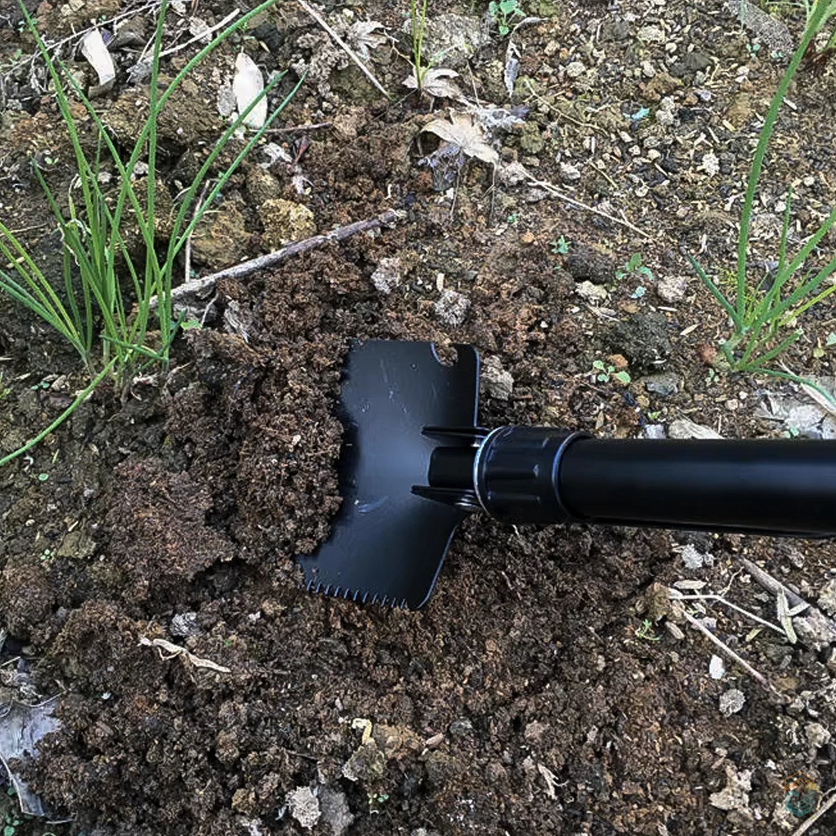 Black tactical camping shovel digging into loose soil, demonstrating serrated blade and sturdy build for outdoor survival, gardening, or campsite preparation