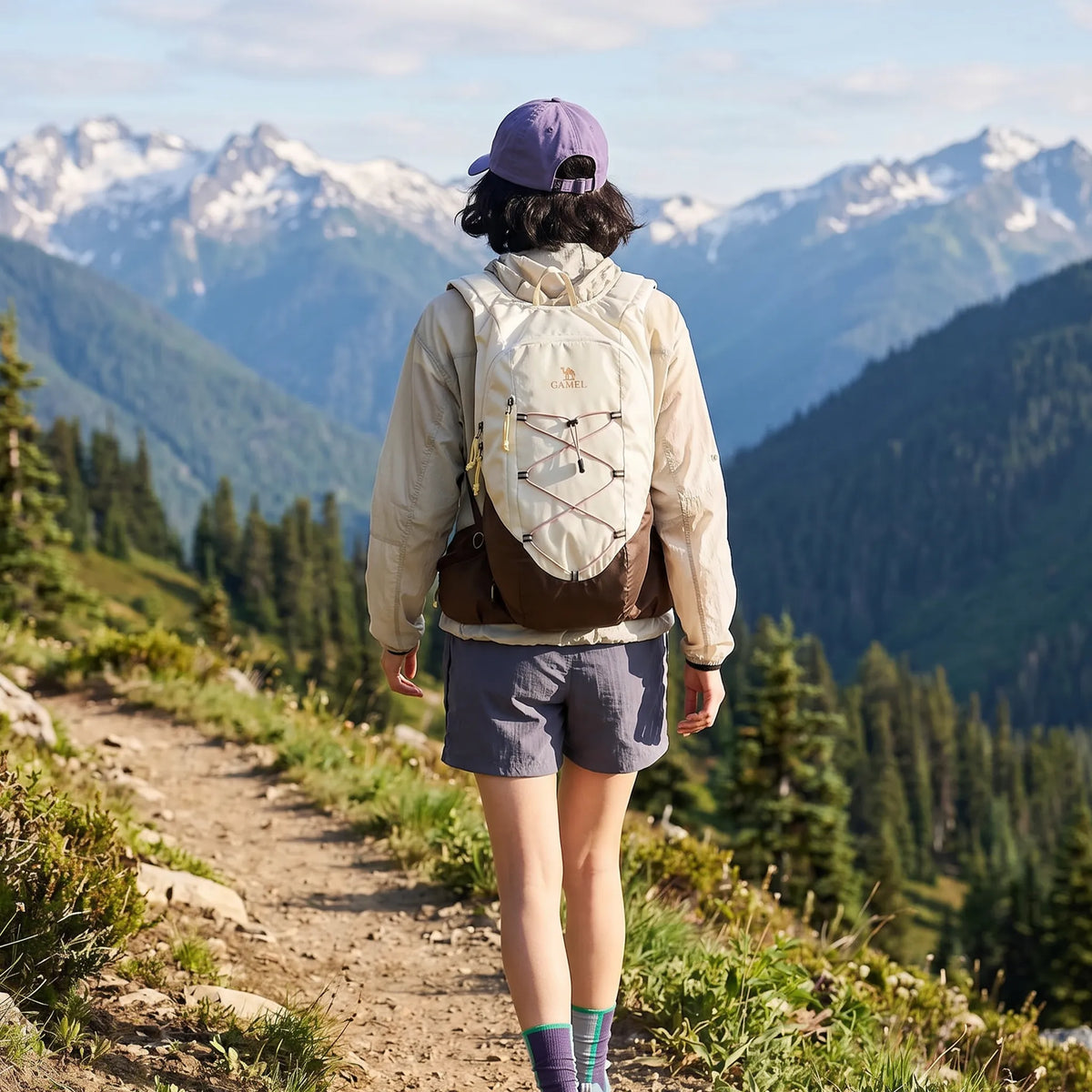 Hiker wearing a brown Camel 20L lightweight hiking backpack while walking along a mountain trail with snow-covered peaks.