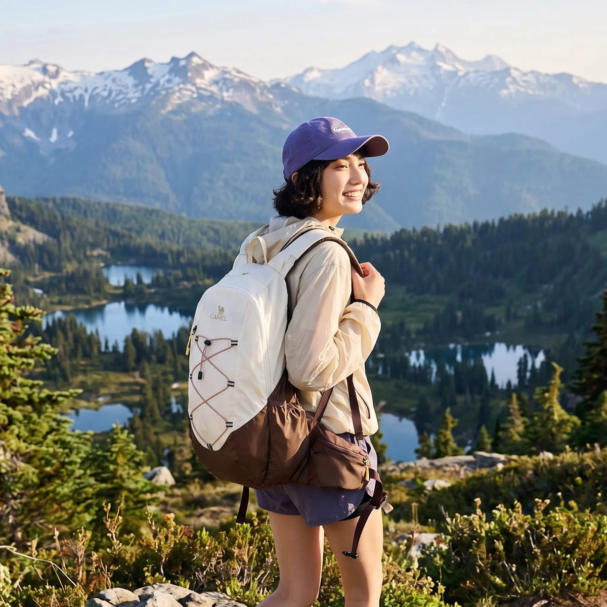 Woman wearing a brown Camel 20L lightweight hiking backpack on a mountain trail overlooking alpine lakes and snow-covered peaks.