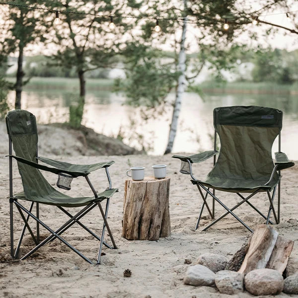 Two green camping chairs set up around a campfire on a sandy shore by a lake, with a log table and mugs, surrounded by trees and string lights.