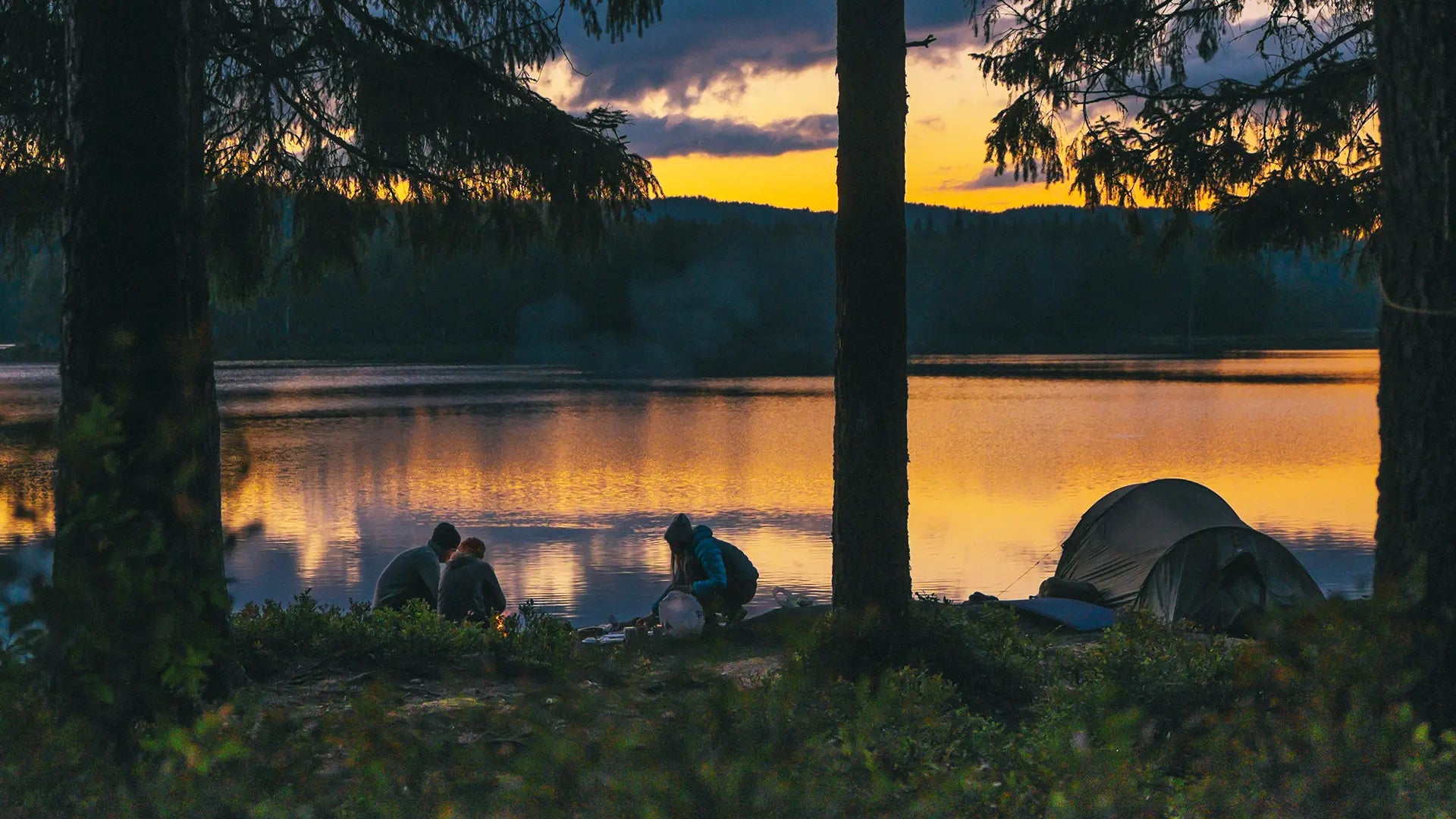 People camping near a lake at sunset, with a tent and a campfire set up under tall trees, reflected in the calm water with a beautiful orange and purple sky.