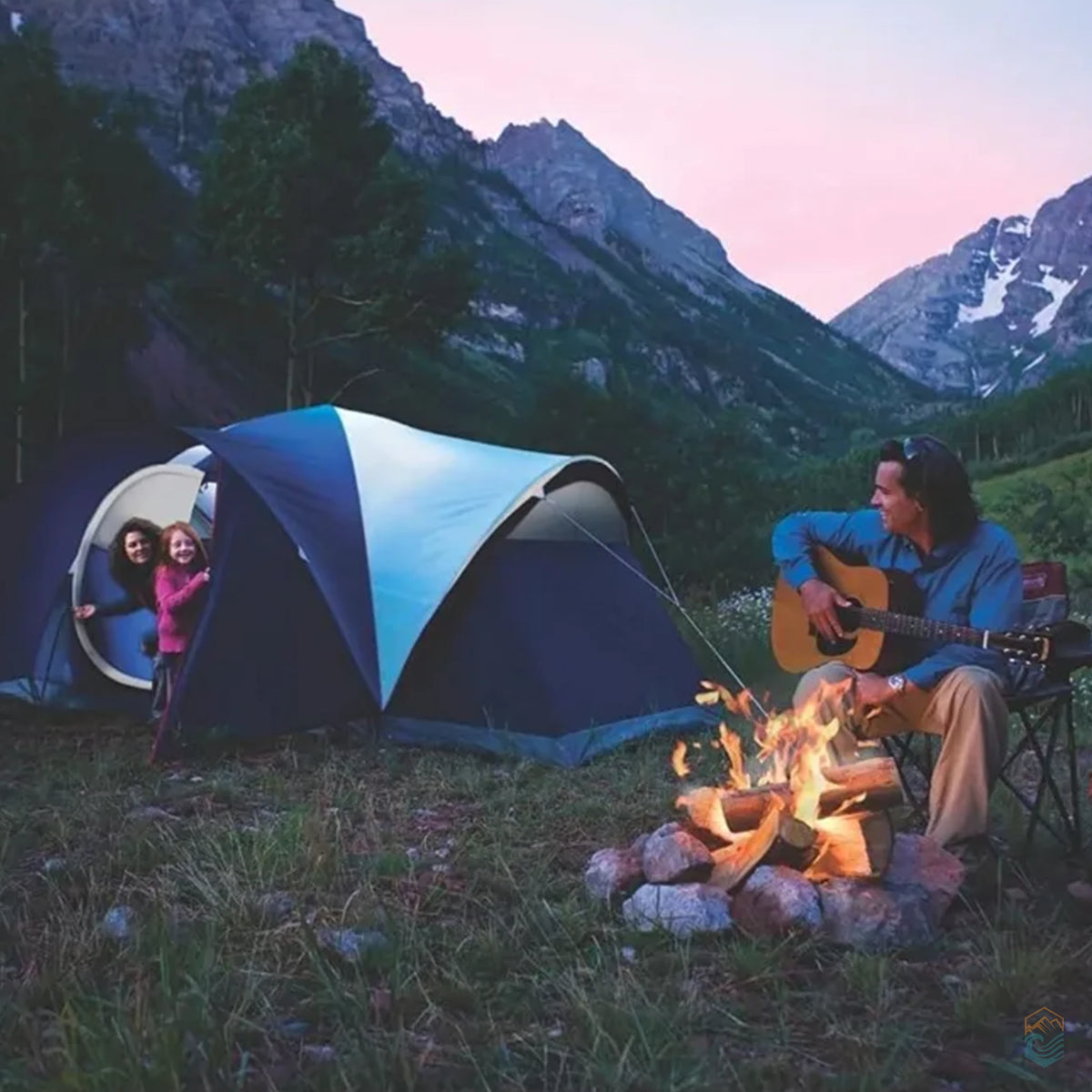 Family camping scene at dusk with a blue Coleman tent set up near a campfire in a mountain valley. A man plays guitar by the fire while two people smile from the tent doorway.