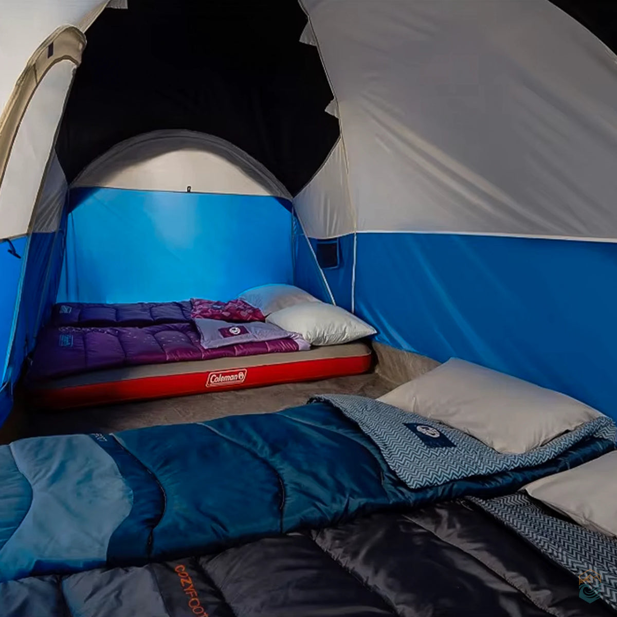 Interior view of a Coleman camping tent with two sleeping areas, including a red Coleman air mattress and multiple sleeping bags and pillows laid out on the tent floor.