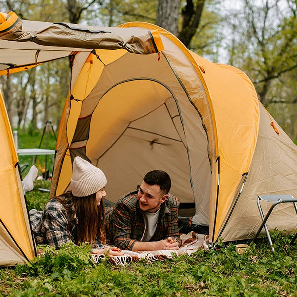 A young couple enjoys a serene camping experience in a lush green forest, lounging outside their yellow tent. The man and woman, clad in casual outdoor wear, engage in a heartfelt conversation amidst the tranquil surroundings, with tall trees and vibrant green grass encapsulating the essence of an intimate nature retreat.