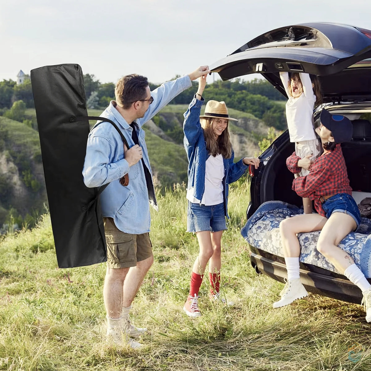 Family enjoying a camping trip near a car, with a man carrying a black folding camping cot bag over his shoulder