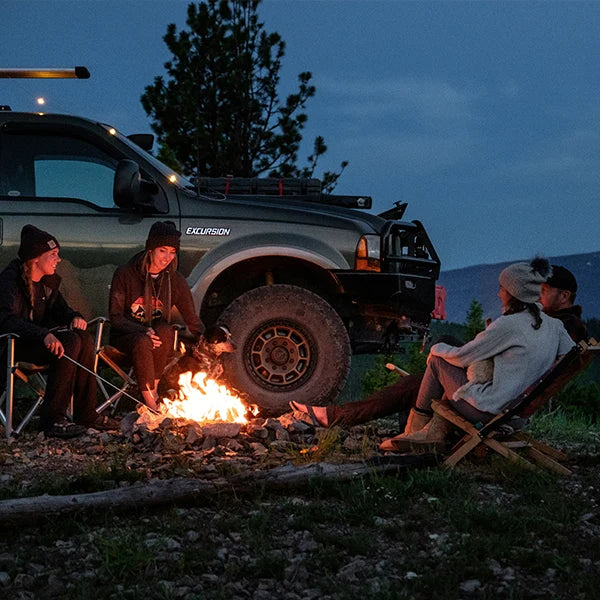 A cozy evening scene with a group of friends and a dog gathered around a campfire in the wilderness, with a rugged vehicle in the background. The warmth of the fire contrasts with the dusk sky and mountainous backdrop, creating a perfect atmosphere for outdoor adventures and bonding. The friends, visibly relaxed and engaged in conversation, enjoy the tranquility and beauty of their remote campsite, showcasing a classic camping experience.