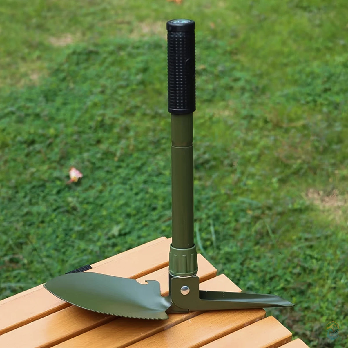 Green folding camping shovel fully assembled and upright on a wooden bench, showcasing integrated pickaxe, serrated blade, and compass grip in a grassy outdoor setting