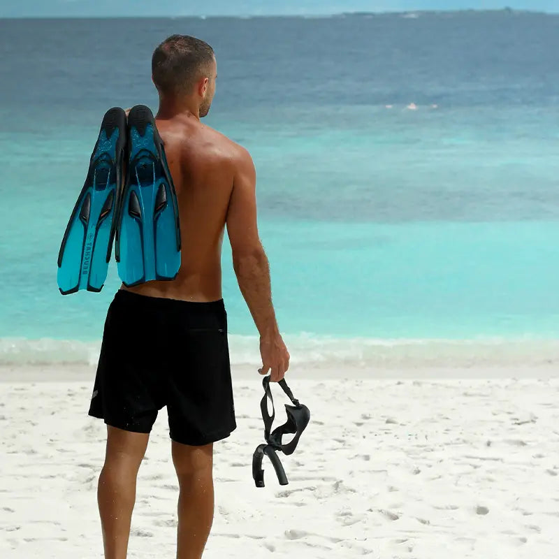 A man standing on a sandy beach, holding a snorkel mask and fins over his shoulder, looking out at the turquoise ocean under a cloudy sky.