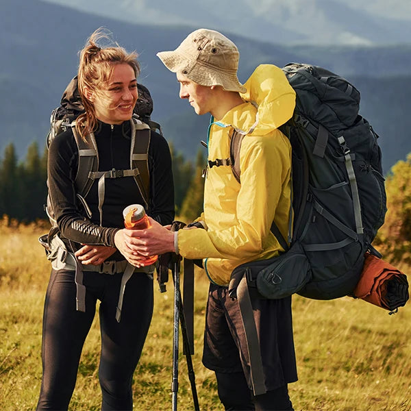 A young couple, fully equipped with heavy backpacks and hiking poles, shares a warm, intimate moment against a stunning backdrop of distant rolling hills and lush greenery. The woman smiles gently at her partner, creating a sense of companionship and affection amidst the breathtaking mountainous landscape, capturing the essence of adventure and togetherness in the great outdoors.