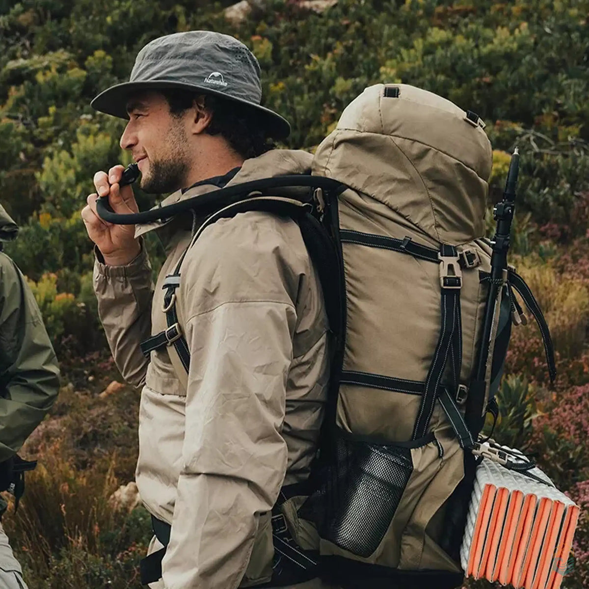 Camper with Naturehike 70L backpack drinking from hydration system, equipped for hiking with poles and foam mat, in a lush outdoor setting.