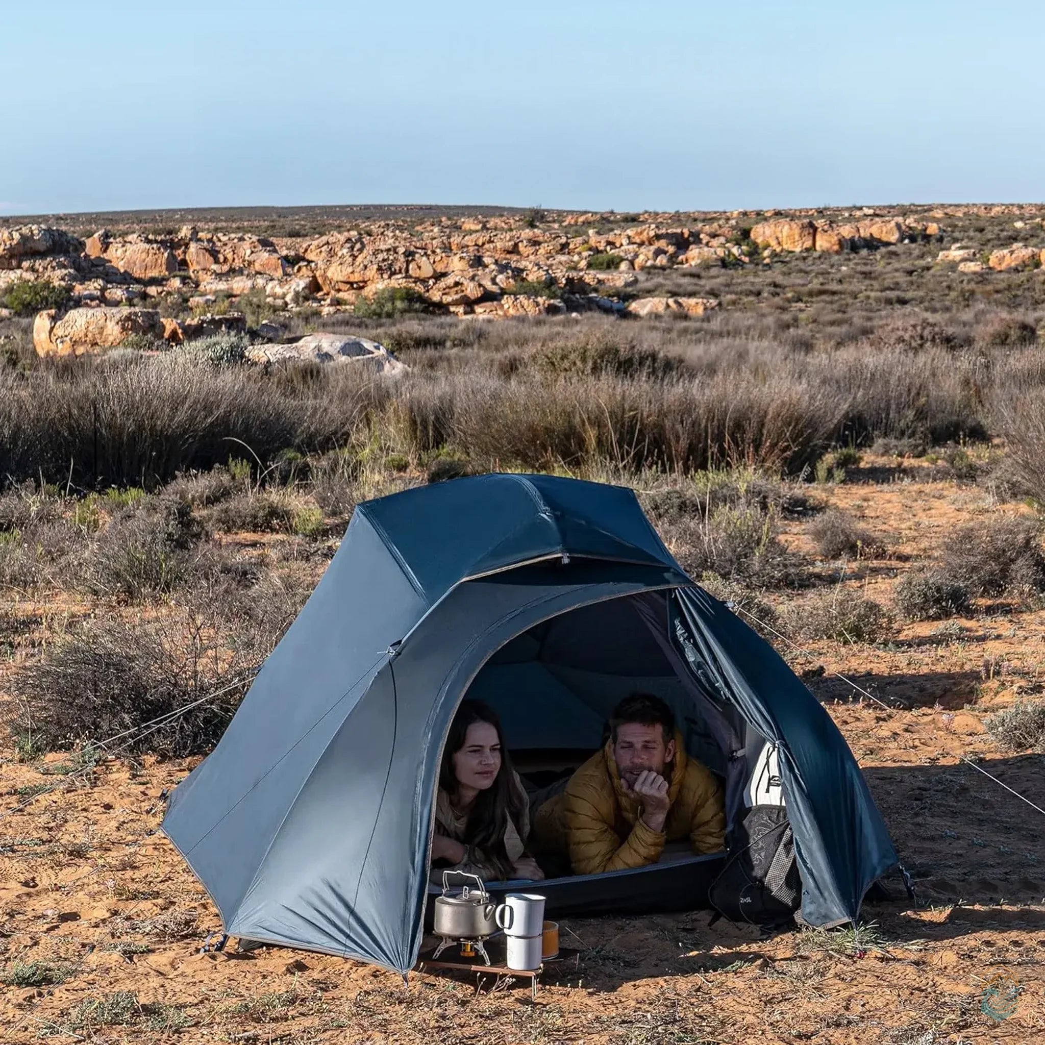 Couple relaxing inside a navy blue Naturehike Cloud-Up 3 Person Tent with the front flap open, set in a dry, rocky outdoor landscape with a portable stove and coffee gear in front