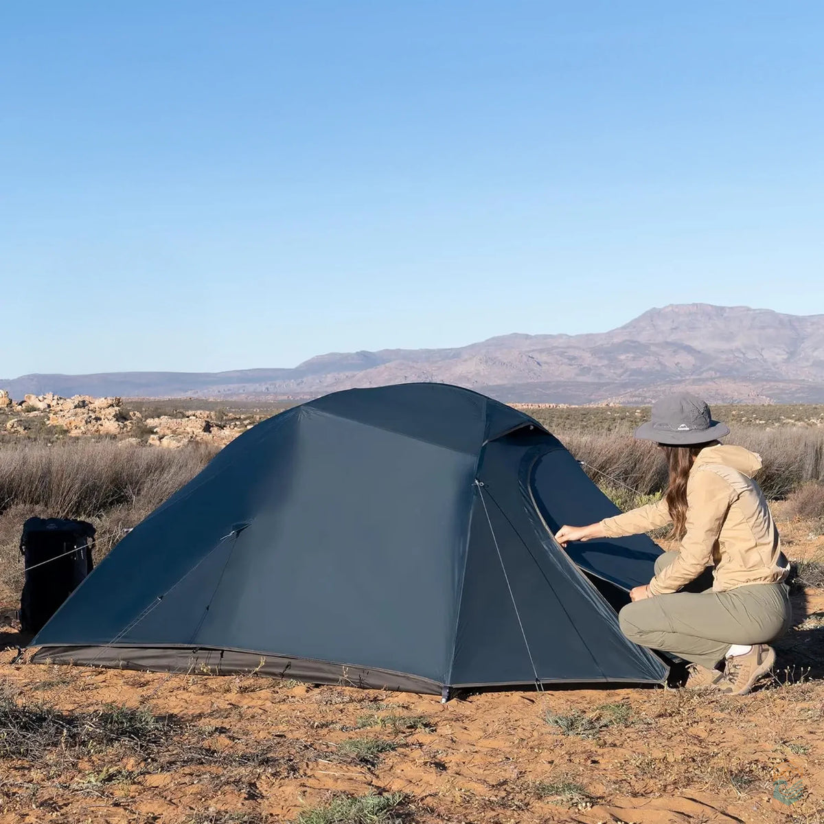 Woman adjusting the entrance of a navy blue Naturehike Cloud-Up 3 Person Tent set up in a desert-like terrain with mountains in the background