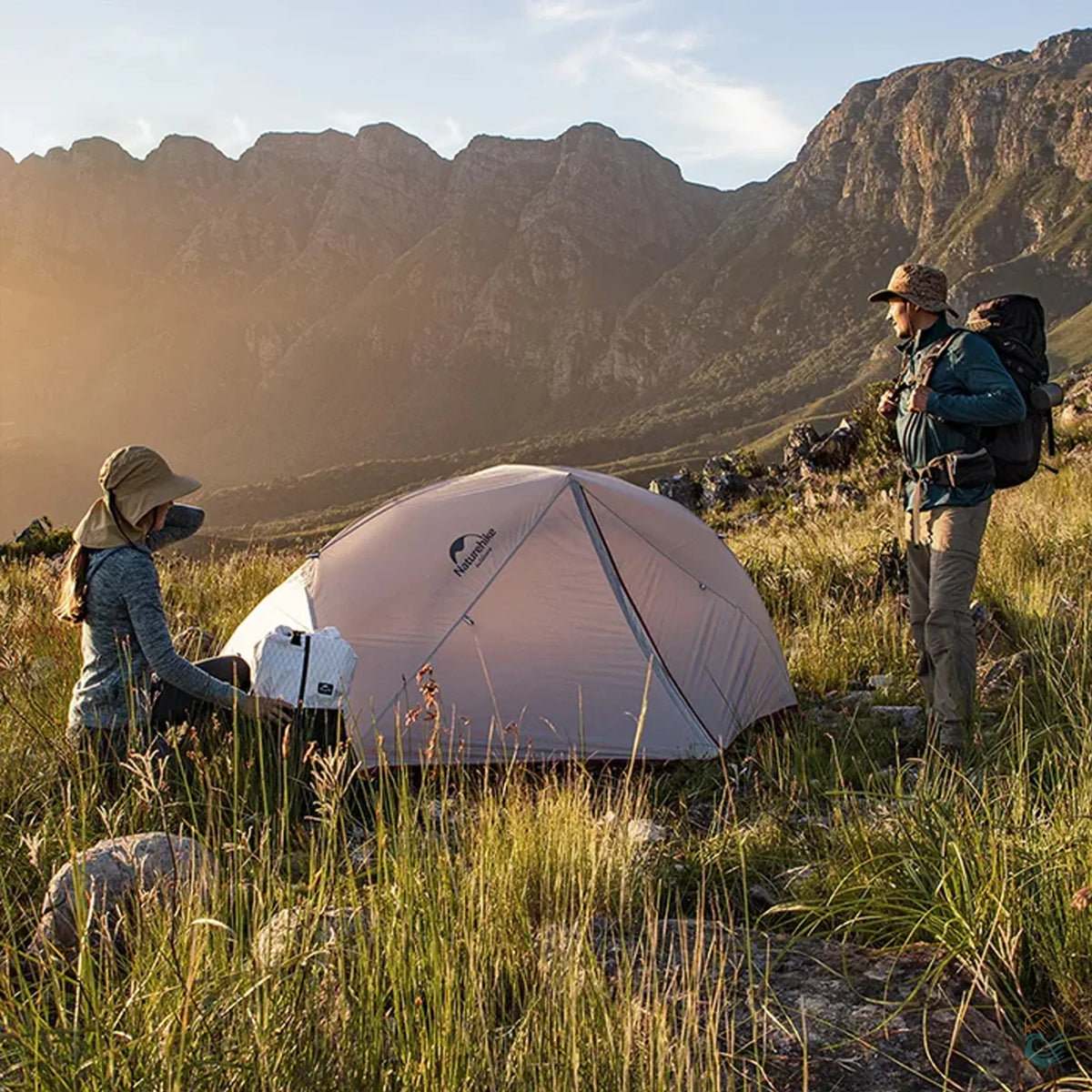 Two hikers setting up a Naturehike Star River 2-person gray tent in a scenic mountain valley during golden hour, surrounded by grass and rugged terrain